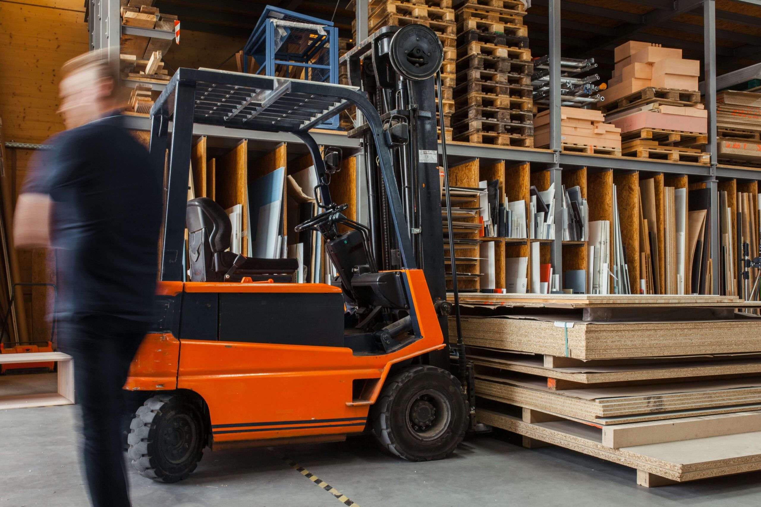 Blurry figure of a worker in a warehouse operating a forklift in a warehouse among stacks of materials and stock, depicting a busy Inventory.